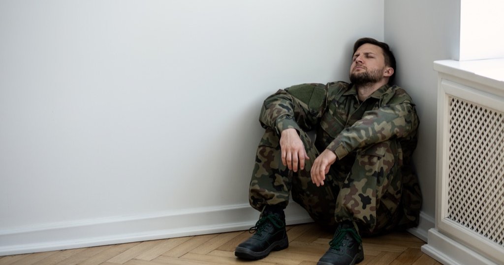 Depressed army man in uniform sitting in a corner of an empty room. Place for your poster on the wall
