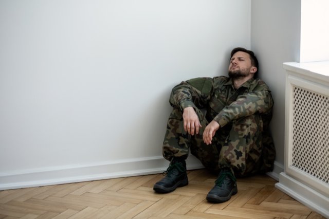 Depressed army man in uniform sitting in a corner of an empty room. Place for your poster on the wall